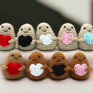 May include: A row of eight crocheted potato-shaped plushies, each holding a different colored heart. The plushies are beige and brown with black eyes and smiling mouths.