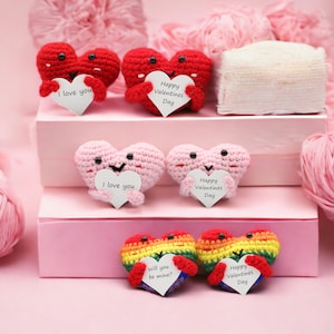 May include: Six crocheted hearts with Valentine's Day messages. The hearts are in various colors, including red, pink, and rainbow. The hearts are arranged on a pink background with pink yarn and a white box.