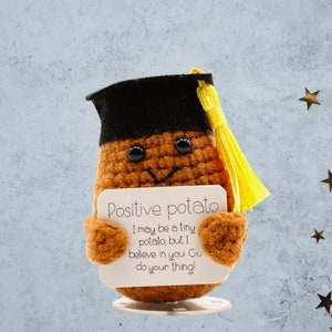 May include: A brown crocheted potato wearing a black graduation cap with a yellow tassel, holding a white card that says "Positive potato I may be a tiny potato, but I believe in you Go do your thing!"