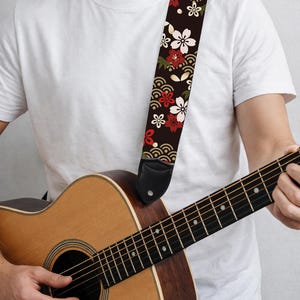 May include: Acoustic guitar with a decorative strap. The strap is black with a floral pattern of white, red, and green flowers. The guitar is made of wood and has a natural finish. The strap is attached to the guitar with black leather ends.