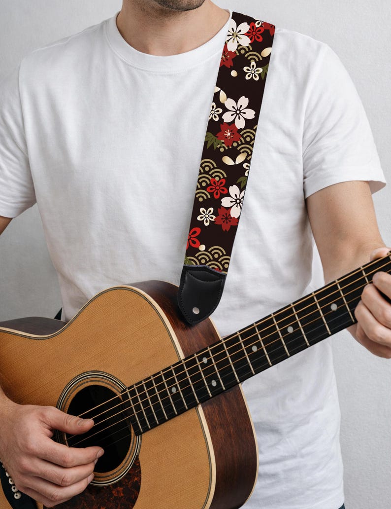 May include: Acoustic guitar with a decorative strap. The strap is black with a floral pattern of white, red, and green flowers. The guitar is made of wood and has a natural finish. The strap is attached to the guitar with black leather ends.