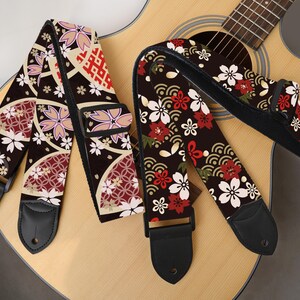 May include: Two guitar straps with Japanese-inspired floral patterns. One strap features a design with white, pink, and red flowers on a dark brown background. The other strap has a similar design with a different arrangement of flowers and patterns.