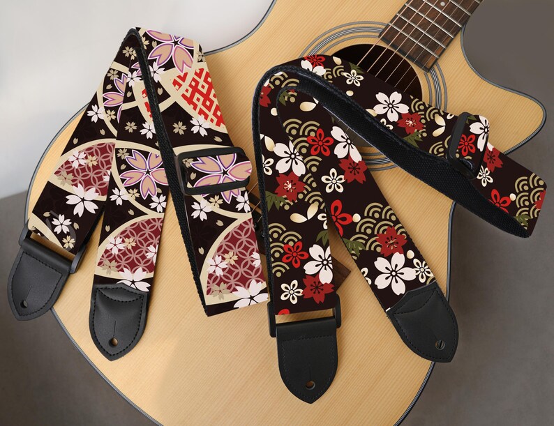May include: Two guitar straps with Japanese-inspired floral patterns. One strap features a design with white, pink, and red flowers on a dark brown background. The other strap has a similar design with a different arrangement of flowers and patterns.