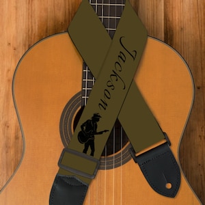 Könnte beinhalten: Ein olivgrüner Gitarrengurt mit dem Namen "Jackson" in schwarzer Schrift und einer Silhouette eines Gitarristen. Der Gurt ist über einer klassischen Akustikgitarre mit Holzfinish drapiert. Schwarze Lederenden vervollständigen das Design.