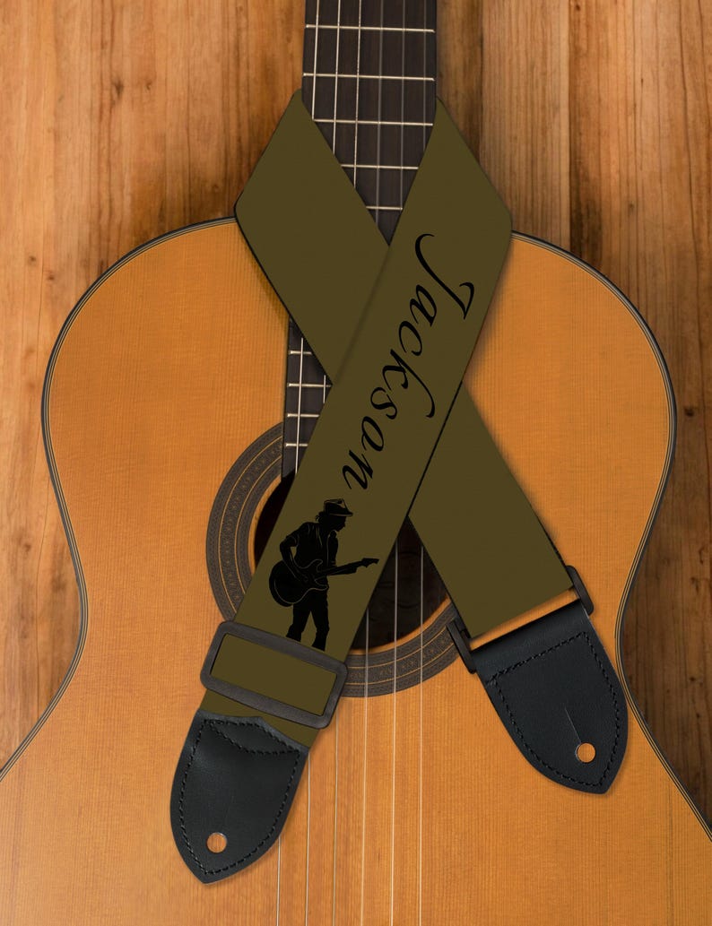 May include: An olive green guitar strap with the name "Jackson" printed in black script. A black silhouette of a guitarist is also featured. The strap is draped over a classical acoustic guitar with a wooden background.