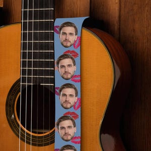 May include: Acoustic guitar with a light blue guitar strap featuring a repeating pattern of a man's face and red lipstick kisses. The guitar has a natural wood finish and is set against a wooden background.
