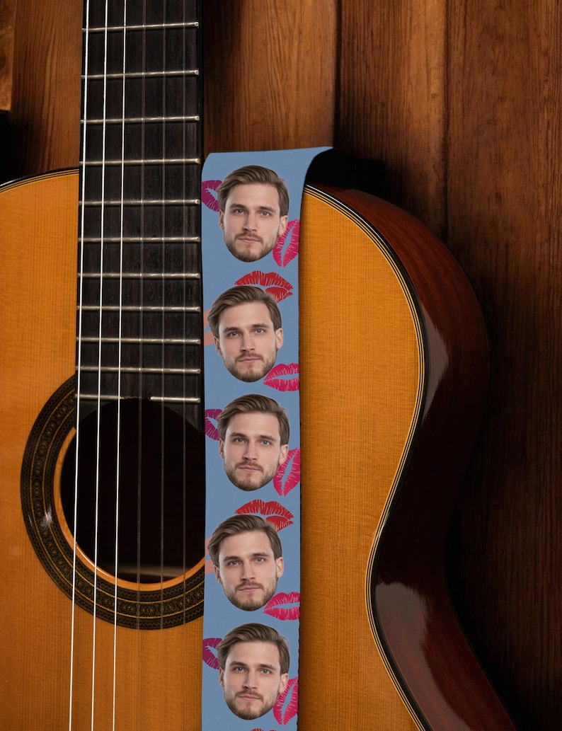 May include: Acoustic guitar with a light blue guitar strap featuring a repeating pattern of a man's face and red lipstick kisses. The guitar has a natural wood finish and is set against a wooden background.