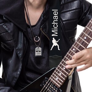 May include: Black guitar strap with the name "Michael" in white lettering and a white silhouette of a guitarist. The strap is attached to a black electric guitar. The image also shows a silver dog tag necklace.