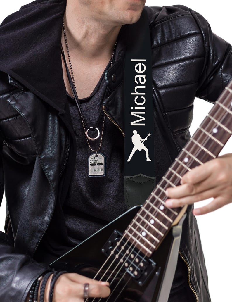 May include: Black guitar strap with the name "Michael" in white lettering and a white silhouette of a guitarist. The strap is attached to a black electric guitar. The image also shows a silver dog tag necklace.