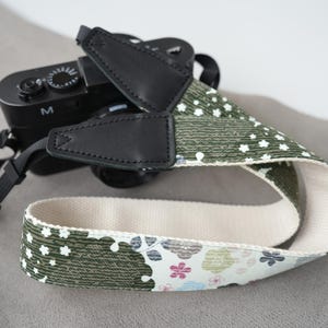 Könnte beinhalten: Ein Kameragurt aus gemustertem Stoff mit olivgrünen Abschnitten, die mit kleinen weißen Blumen verziert sind, und einem weißen Abschnitt mit bunten Blumenmotiven. Der Gurt verfügt über schwarze Lederbefestigungen und schwarzes Nylongewebe.