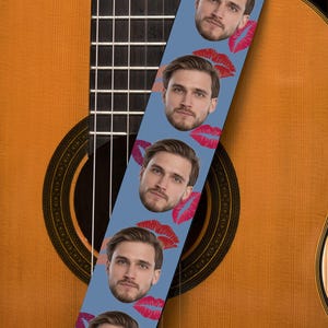 May include: A light blue guitar strap featuring a repeating pattern of a man's face and red lips. The strap is positioned over a wooden acoustic guitar, showcasing its sound hole and strings.