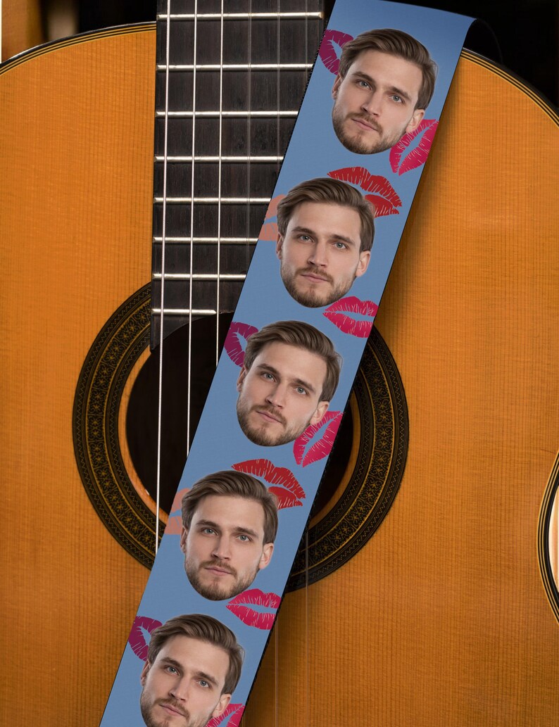 May include: A light blue guitar strap featuring a repeating pattern of a man's face and red lips. The strap is positioned over a wooden acoustic guitar, showcasing its sound hole and strings.