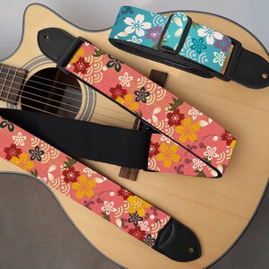 May include: Three guitar straps with floral patterns rest on an acoustic guitar. The straps feature a pink background with yellow, red, and white flowers. One strap is blue with white flowers. The guitar is light brown.