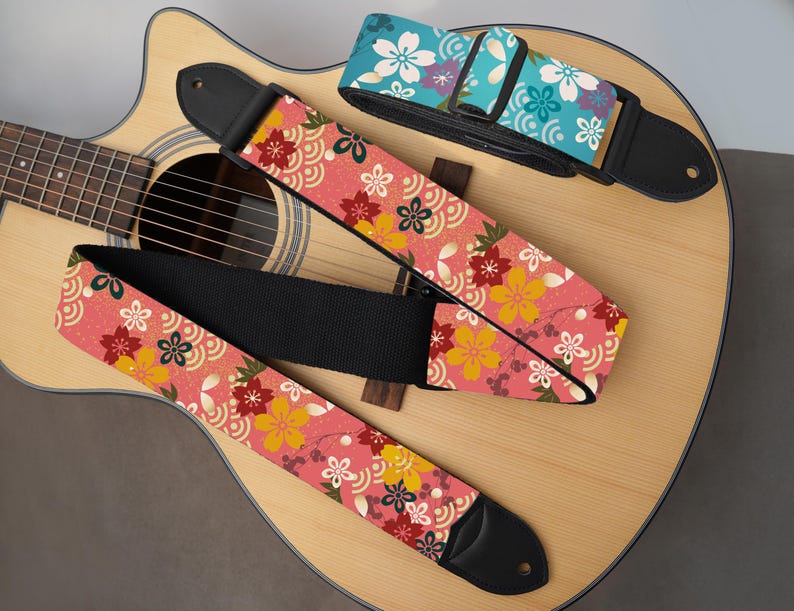 May include: Three guitar straps with floral patterns rest on an acoustic guitar. The straps feature a pink background with yellow, red, and white flowers. One strap is blue with white flowers. The guitar is light brown.