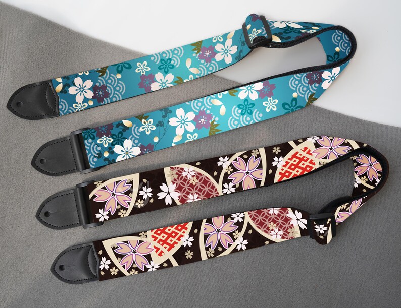 May include: Three guitar straps with floral patterns. One strap is teal with white and purple flowers and a wave design. The other two straps are brown with pink and white flowers and geometric shapes. All straps have black leather ends.