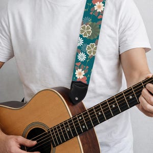 May include: Acoustic guitar with a teal floral guitar strap. The strap features a repeating pattern of flowers in shades of white, coral, and olive green. The guitar is made of wood and has a natural finish.
