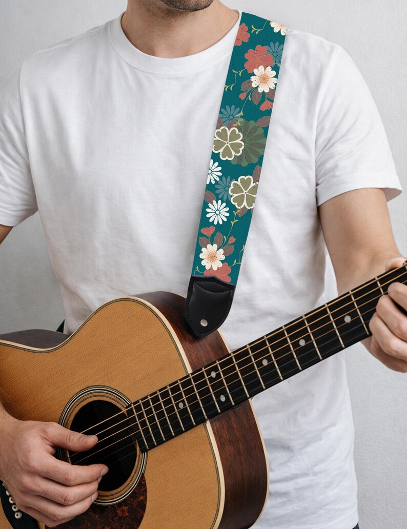May include: Acoustic guitar with a teal floral guitar strap. The strap features a repeating pattern of flowers in shades of white, coral, and olive green. The guitar is made of wood and has a natural finish.
