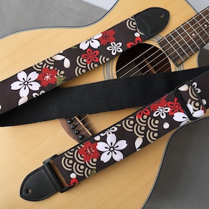 May include: Acoustic guitar with a floral patterned guitar strap. The strap features a dark brown background with white and red flower designs. The strap is made of fabric and has black leather ends.