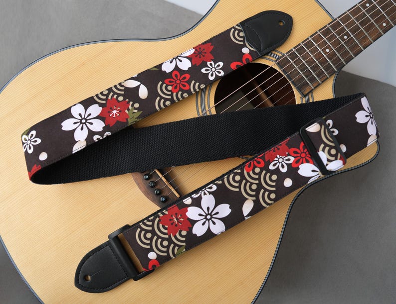 May include: Acoustic guitar with a floral patterned guitar strap. The strap features a dark brown background with white and red flower designs. The strap is made of fabric and has black leather ends.