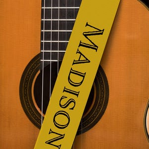 May include: Acoustic guitar with a yellow strap. The strap is printed with the name "MADISON" in black capital letters. The guitar has a wooden body and a black fretboard.