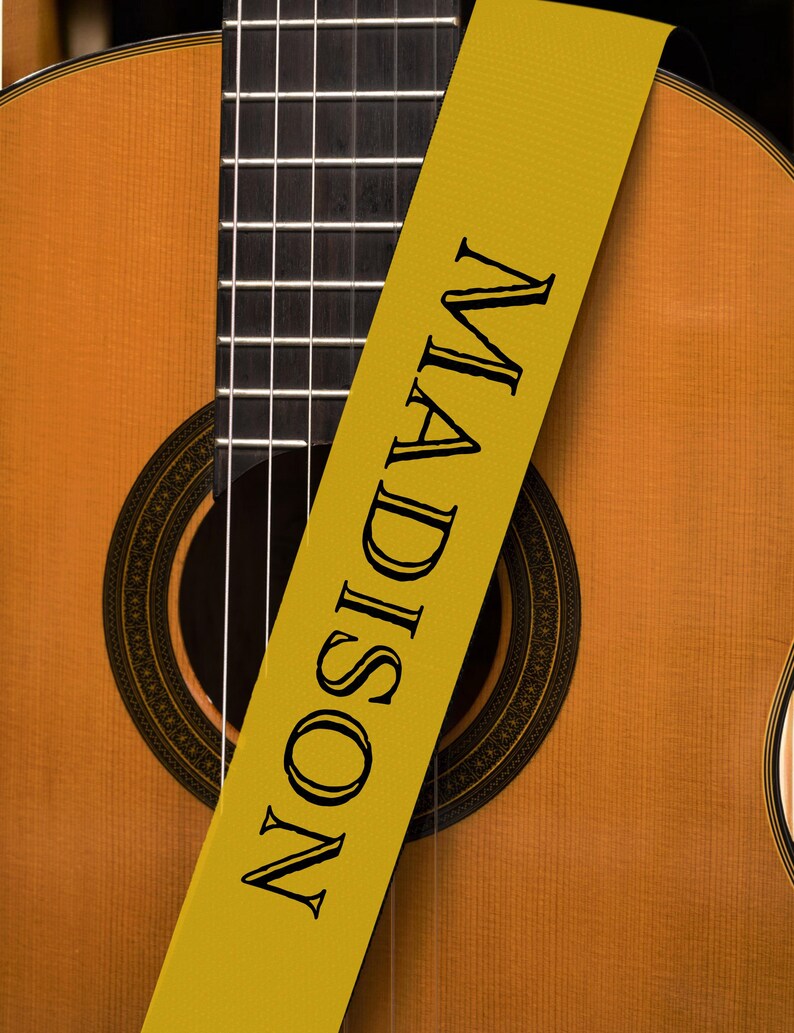 May include: Acoustic guitar with a yellow strap. The strap is printed with the name "MADISON" in black capital letters. The guitar has a wooden body and a black fretboard.