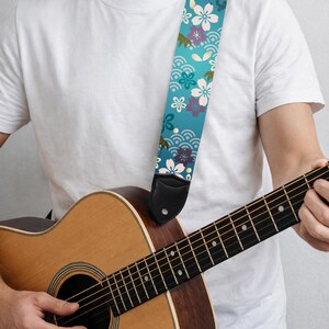 May include: A guitar strap with a teal background and a floral pattern in white, purple, and green. The strap is made of fabric and has black leather ends. The guitar is a natural wood color.