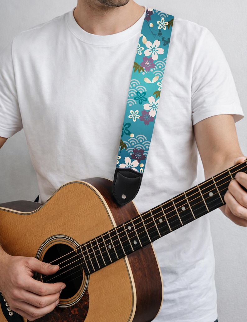 May include: A guitar strap with a teal background and a floral pattern in white, purple, and green. The strap is made of fabric and has black leather ends. The guitar is a natural wood color.