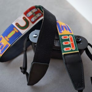 May include: A camera strap featuring a colorful pattern of stylized letters and shapes in red, blue, yellow, and green on a white background. The strap includes black woven fabric sections, black leather-like end tabs, and adjustable black plastic buckles.