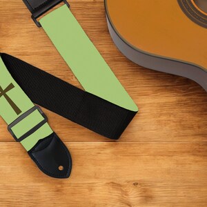 May include: A guitar strap with a light green background and a brown cross design. The strap has a black woven section and a black leather end piece. A brown acoustic guitar is partially visible in the background.