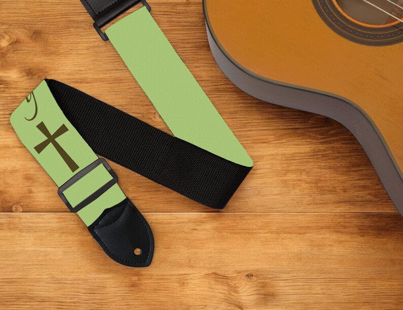 May include: A guitar strap with a light green background and a brown cross design. The strap has a black woven section and a black leather end piece. A brown acoustic guitar is partially visible in the background.