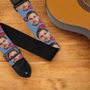 May include: A guitar strap with a light blue background featuring a repeating pattern of a man's face and red lipstick kisses. The strap has a black adjustable portion and a black leather end.