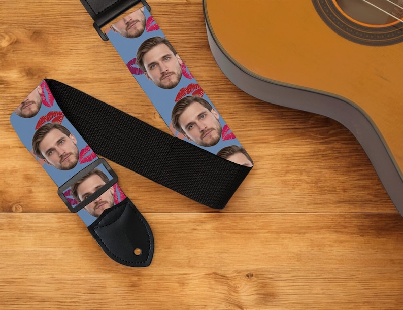 May include: A guitar strap with a light blue background featuring a repeating pattern of a man's face and red lipstick kisses. The strap has a black adjustable portion and a black leather end.