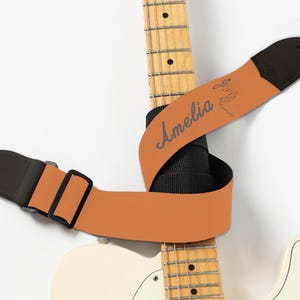 May include: A guitar strap in a warm orange hue, with the name "Amelia" in a script font. The strap has black leather ends and a black adjustable buckle. The strap is wrapped around a white electric guitar.