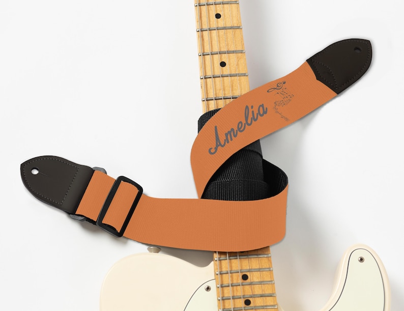 May include: A guitar strap in a warm orange hue, with the name "Amelia" in a script font. The strap has black leather ends and a black adjustable buckle. The strap is wrapped around a white electric guitar.