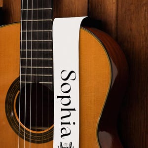 May include: Acoustic guitar with a light brown body and a white guitar strap. The strap features the name "Sophia" in black, along with a rock and roll hand symbol with wings and a skull.