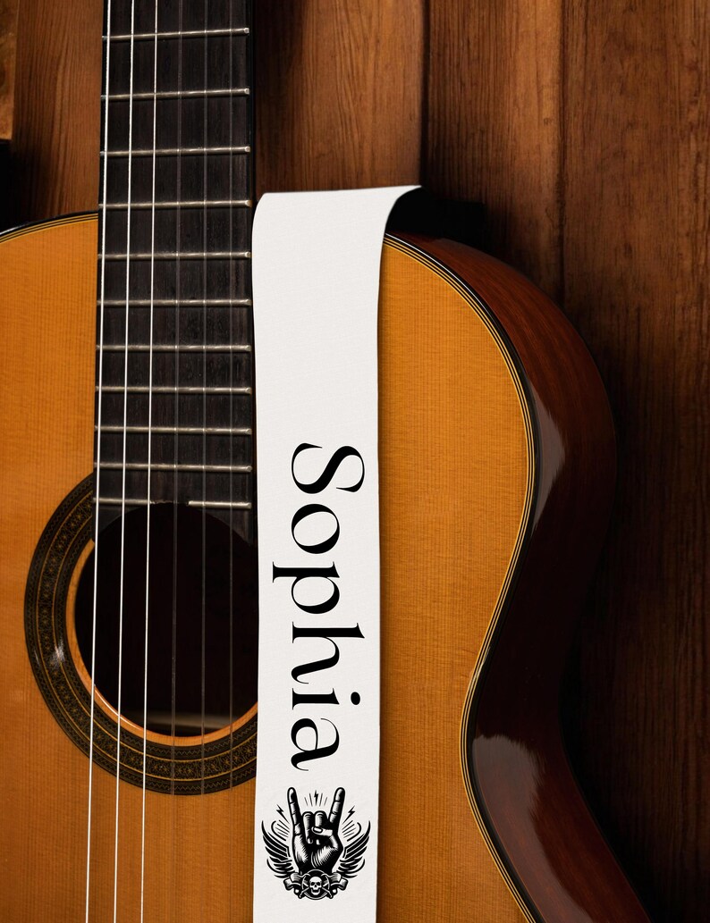 May include: Acoustic guitar with a light brown body and a white guitar strap. The strap features the name "Sophia" in black, along with a rock and roll hand symbol with wings and a skull.