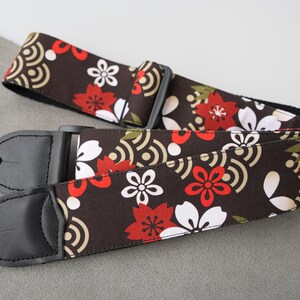 May include: A guitar strap with a floral pattern in red, white, and green on a dark brown background. The strap has black leather ends and a black plastic adjustable buckle. The design includes stylized flowers and wave patterns.