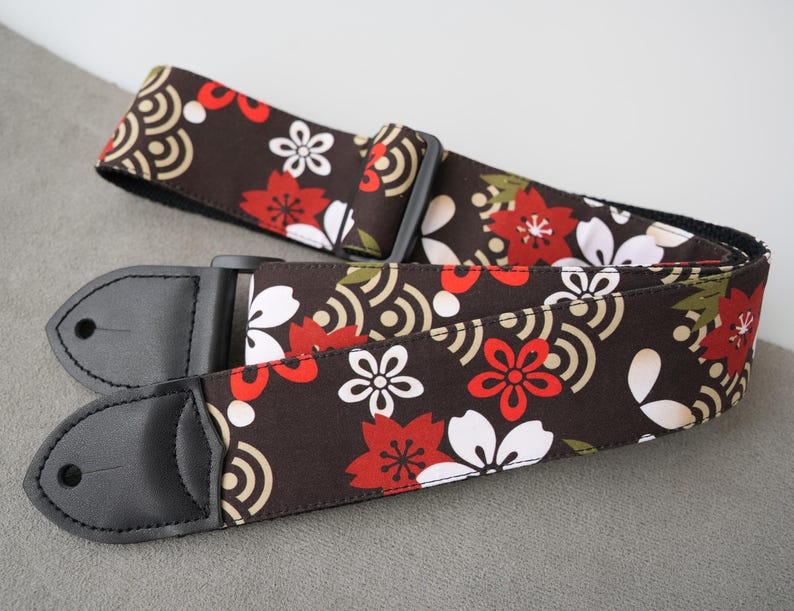May include: A guitar strap with a floral pattern in red, white, and green on a dark brown background. The strap has black leather ends and a black plastic adjustable buckle. The design includes stylized flowers and wave patterns.