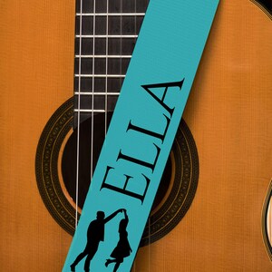 May include: Acoustic guitar with a light brown wooden body and a black fretboard. A teal guitar strap with the name "ELLA" in black lettering and a silhouette of a dancing couple is draped across the guitar.