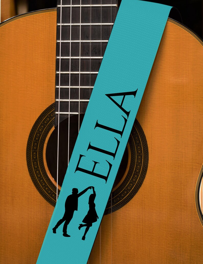 May include: Acoustic guitar with a light brown wooden body and a black fretboard. A teal guitar strap with the name "ELLA" in black lettering and a silhouette of a dancing couple is draped across the guitar.