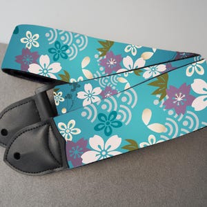 May include: A guitar strap with a teal background and a floral pattern featuring white, purple, and teal flowers. The strap has black leather ends.