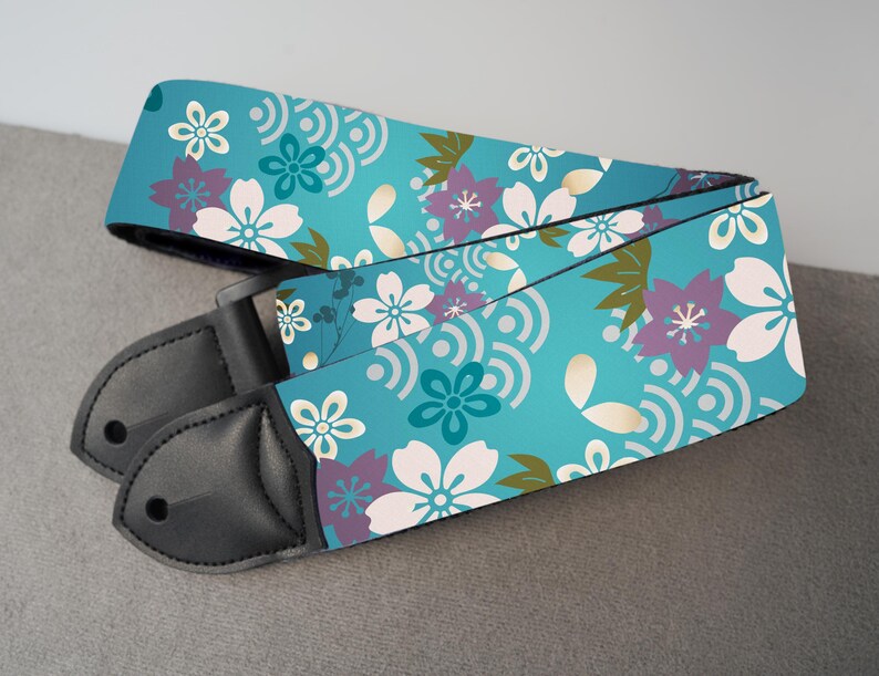 May include: A guitar strap with a teal background and a floral pattern featuring white, purple, and teal flowers. The strap has black leather ends.