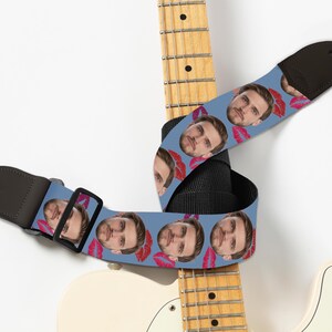 May include: A blue guitar strap featuring a repeating pattern of a person's face and red lipstick kisses. The strap is black on the back and has a black buckle and leather ends. The strap is wrapped around a white and yellow electric guitar.