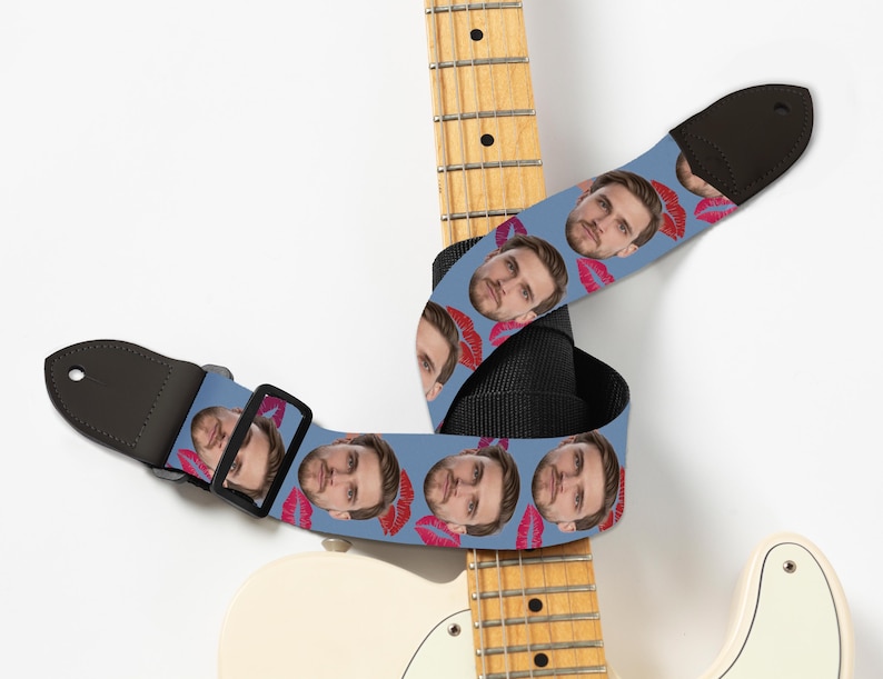 May include: A blue guitar strap featuring a repeating pattern of a person's face and red lipstick kisses. The strap is black on the back and has a black buckle and leather ends. The strap is wrapped around a white and yellow electric guitar.