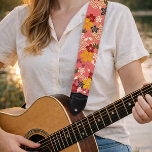 May include: Acoustic guitar with a floral patterned strap. The strap features a pink background with colorful flowers and wave patterns. The guitar is made of wood and has a black leather end piece on the strap.