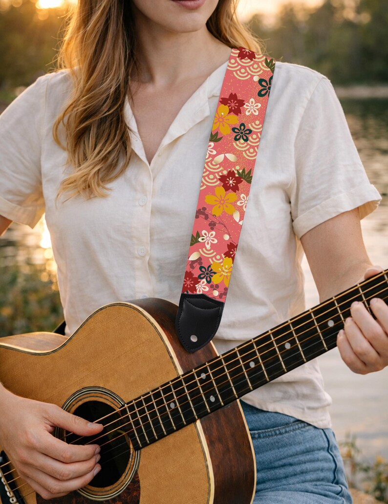 May include: Acoustic guitar with a floral patterned strap. The strap features a pink background with colorful flowers and wave patterns. The guitar is made of wood and has a black leather end piece on the strap.