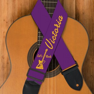 May include: A purple guitar strap with the name "Victoria" in gold script. The strap also features a gold guitar graphic and the words "Let's Rock." Black leather ends and a black adjustable buckle are visible. The strap is draped over a wooden acoustic guitar.