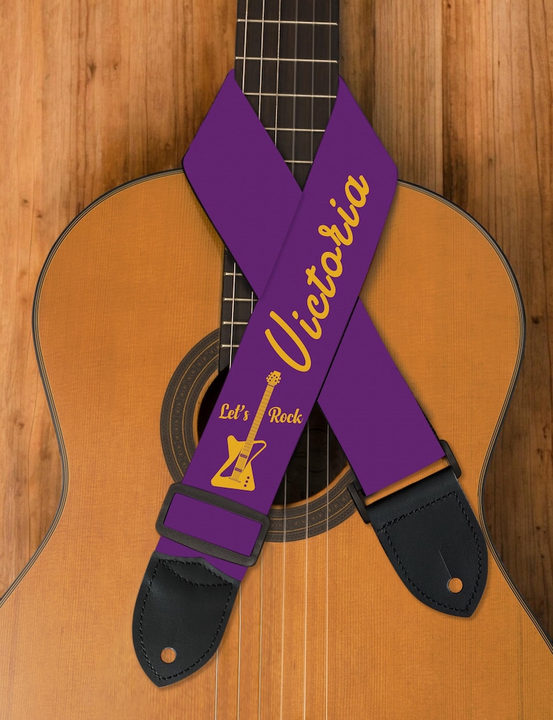 May include: A purple guitar strap with the name "Victoria" in gold script. The strap also features a gold guitar graphic and the words "Let's Rock." Black leather ends and a black adjustable buckle are visible. The strap is draped over a wooden acoustic guitar.