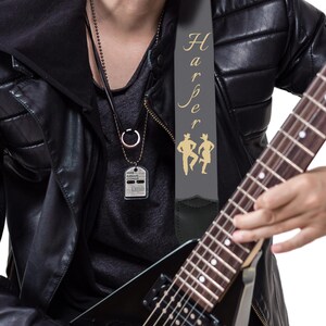 May include: A black electric guitar with a gray guitar strap featuring the name "Harper" in gold script and two gold silhouette figures. The person is wearing a black leather jacket and a silver dog tag necklace.