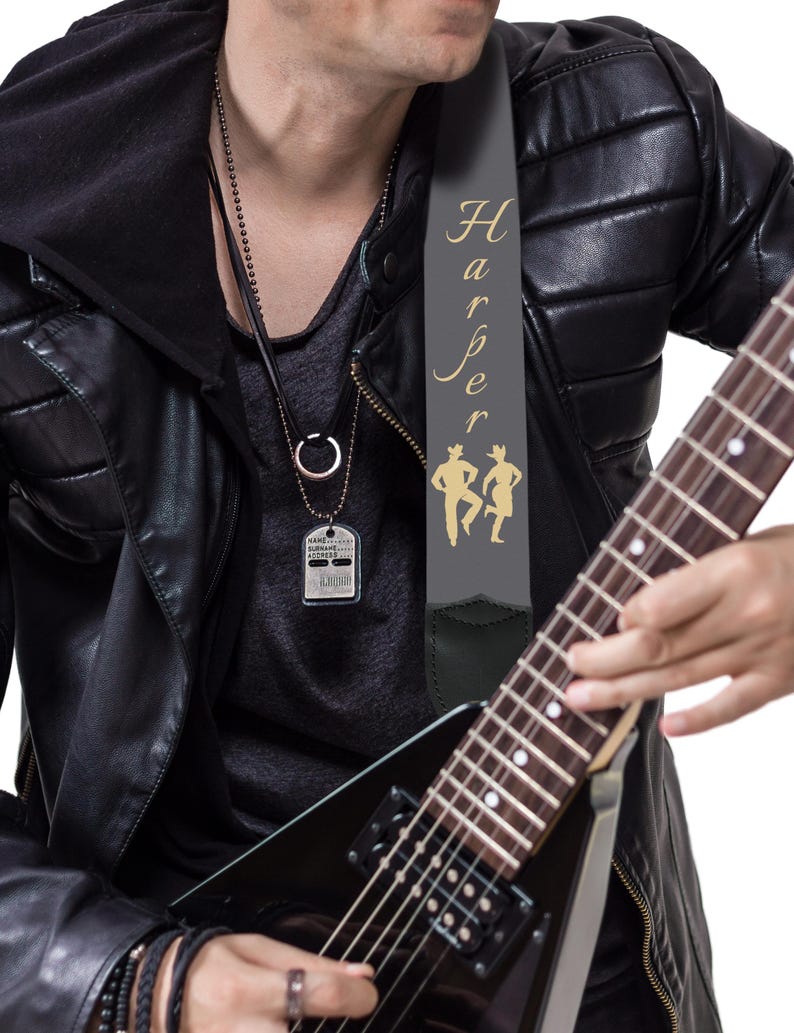 May include: A black electric guitar with a gray guitar strap featuring the name "Harper" in gold script and two gold silhouette figures. The person is wearing a black leather jacket and a silver dog tag necklace.